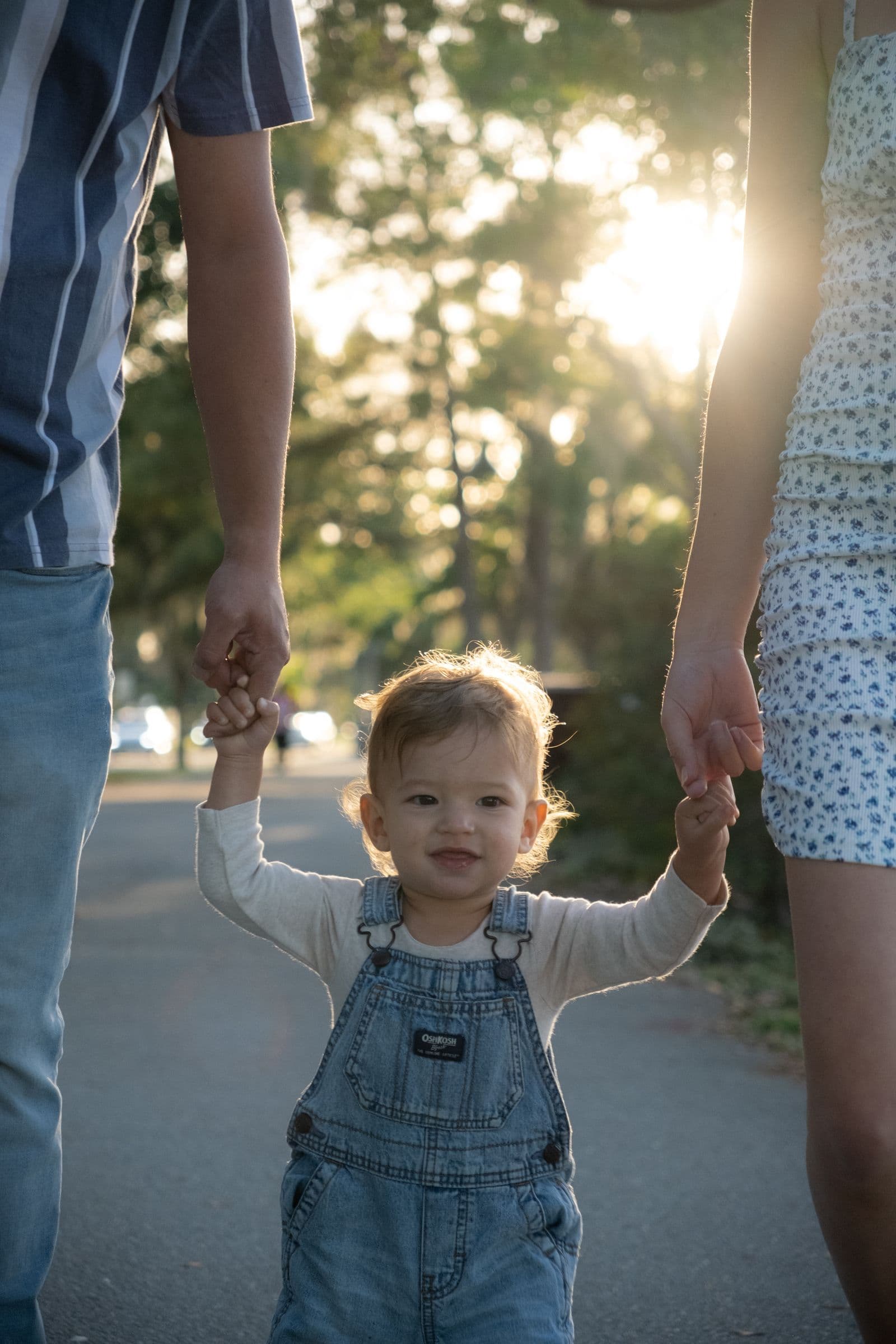 A toddler walking in the park holding both parents' hands