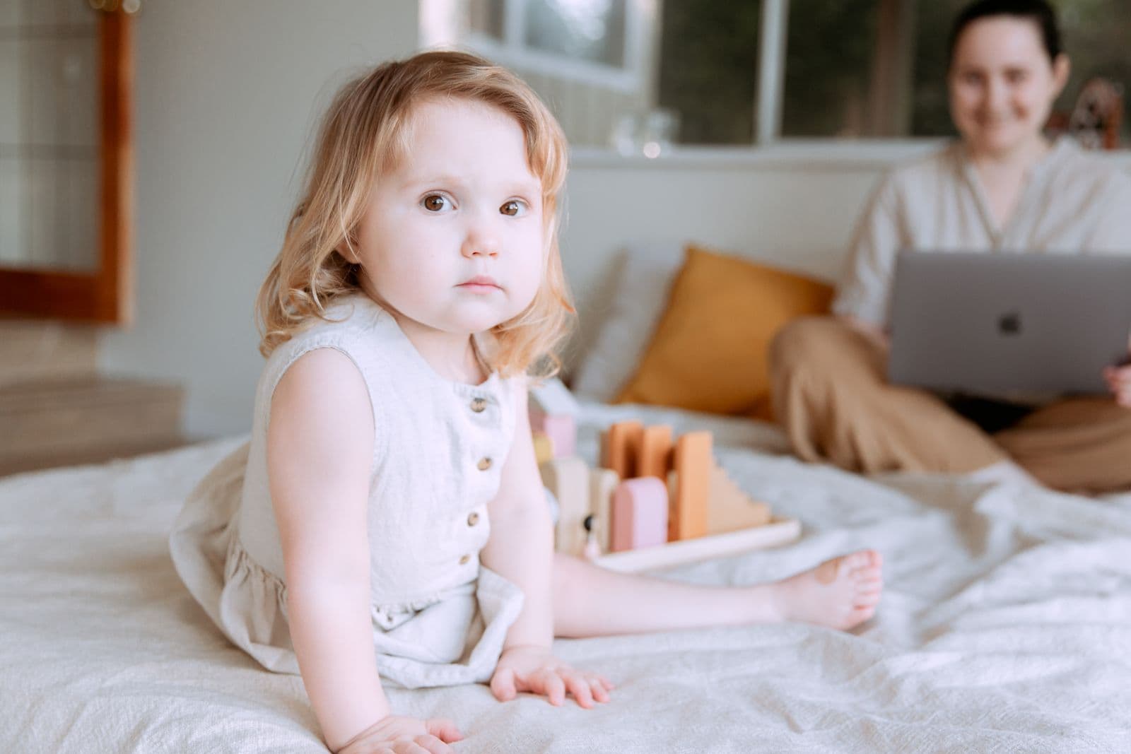 A toddler playing on the bed while mum works on a laptop